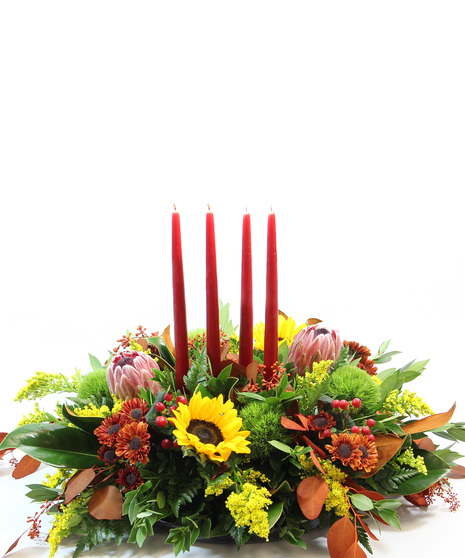 Four 15-inch tapered candles casts a glow over yellow sunflowers, red protea, magnolia leaves, tinted eucalyptus, red hypericum, moss carnations and bronze Viking poms.  
