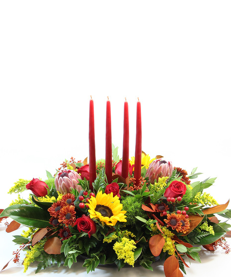 Four 15-inch tapered candles casts a glow over yellow sunflowers, red protea, magnolia leaves, tinted eucalyptus, red hypericum, moss carnations and bronze Viking poms.  