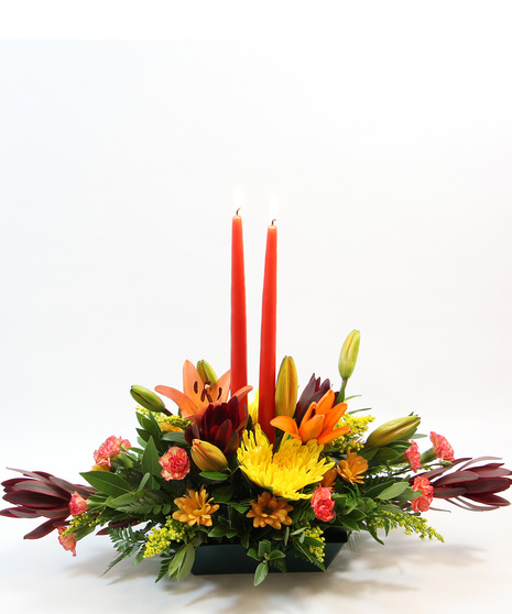 This charming centerpiece showcases two tapered candles surrounded by vibrant orange Asiatic lilies, yellow spider mums, leucadendron, bronze daisies, orange pixie carnations.