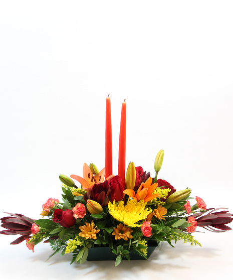 This charming centerpiece showcases two tapered candles surrounded by vibrant orange Asiatic lilies, yellow spider mums, leucadendron, bronze daisies, orange pixie carnations.