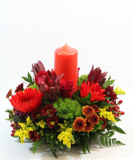 The Fall Grandeur centerpiece features a pillar candle surrounded by vibrant red sunflowers, moss carnations, hypericum, leucadendron, and red daisies. 