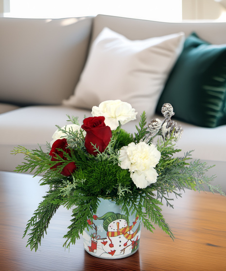Holiday bouquet in whimsical snowman tin with evergreens, white & moss carnations, and frosted pinecones. A cheerful winter gift.
