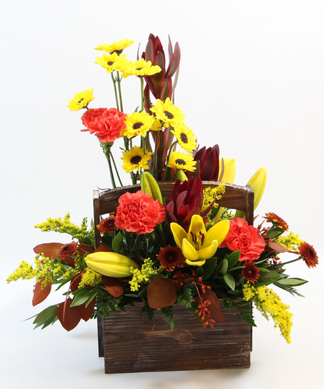 a beautifully crafted bouquet arranged in a wooden basket. Featuring yellow Asiatic lilies, orange carnations, leucadendron, Viking poms, tinted eucalyptus, and fall buttons