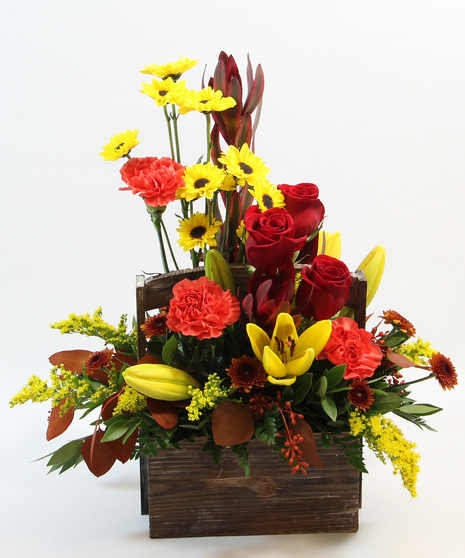 a beautifully crafted bouquet arranged in a wooden basket. Featuring yellow Asiatic lilies, orange carnations, leucadendron, Viking poms, tinted eucalyptus, and fall buttons