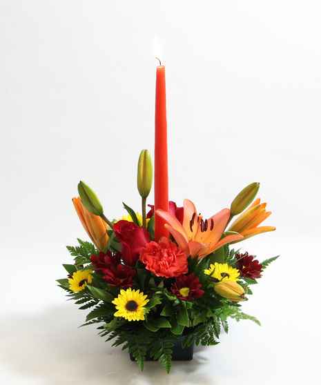 This centerpiece features a single tapered candle surrounded by orange Asiatic lilies, orange carnations, yellow Viking poms, and red daisies.