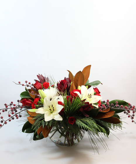 Magnolia leaves and fresh evergreens with white Asiatic lilies, red roses, white spider mums, red pixie carnations, leucadendron, and red berries in a clear glass bubble bowl.