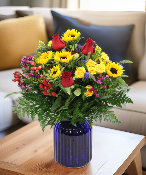 Sunflowers, spray roses, spring asters, and accents in a cobalt blue glass vase. A warm and generous bouquet bursting with color